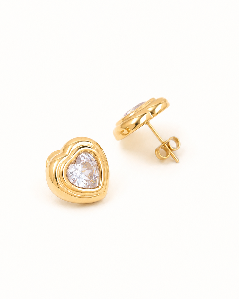 A pair of gold heart stud earrings with a clear heart-shaped gemstone in the center, shown on a plain white background with one earring facing forward and the other angled to show the post and backing.